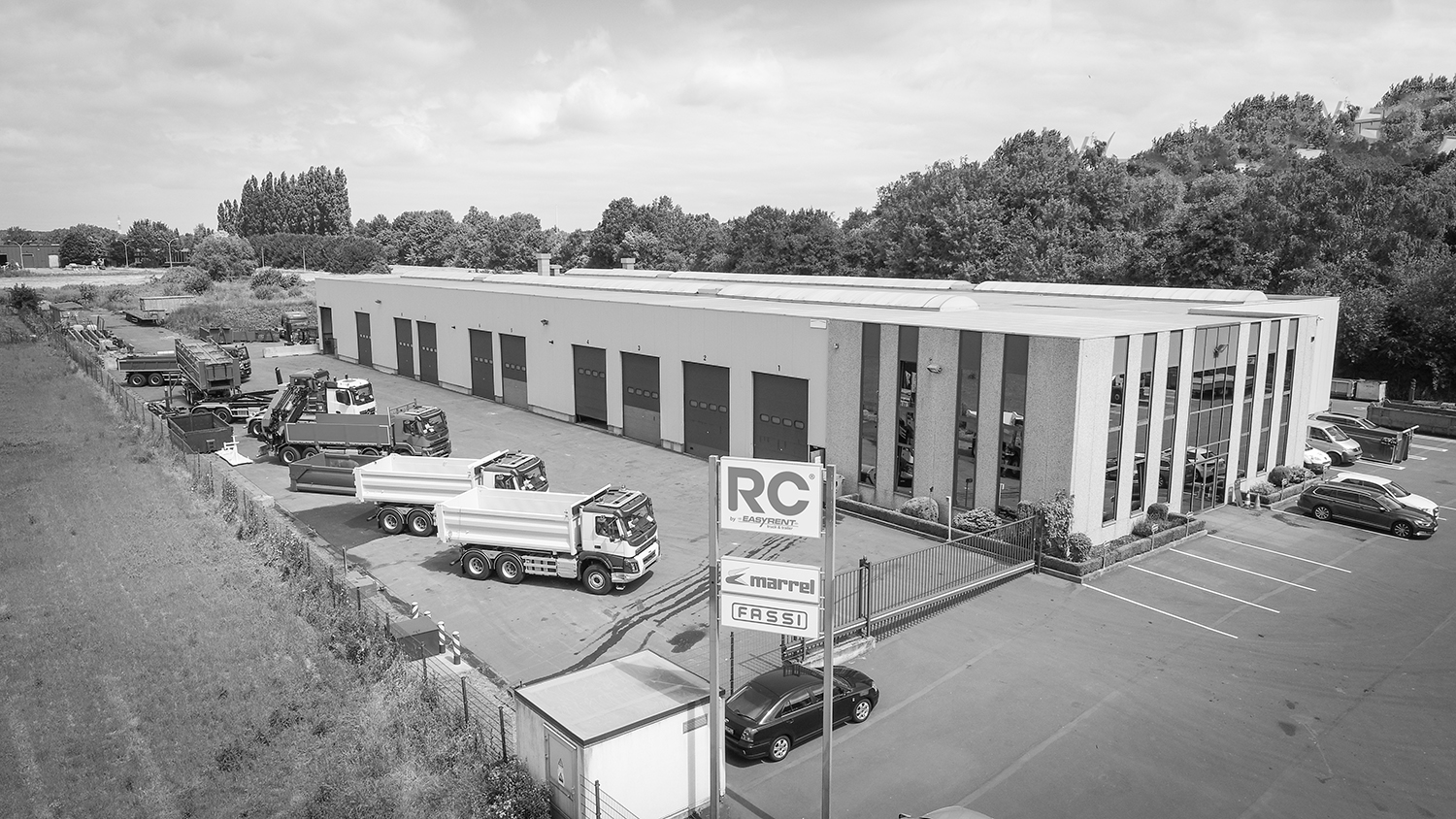 Vue aérienne du site Atelier RC à Herstal, Belgique. Grand hall industriel avec des camions-bennes dans la cour.