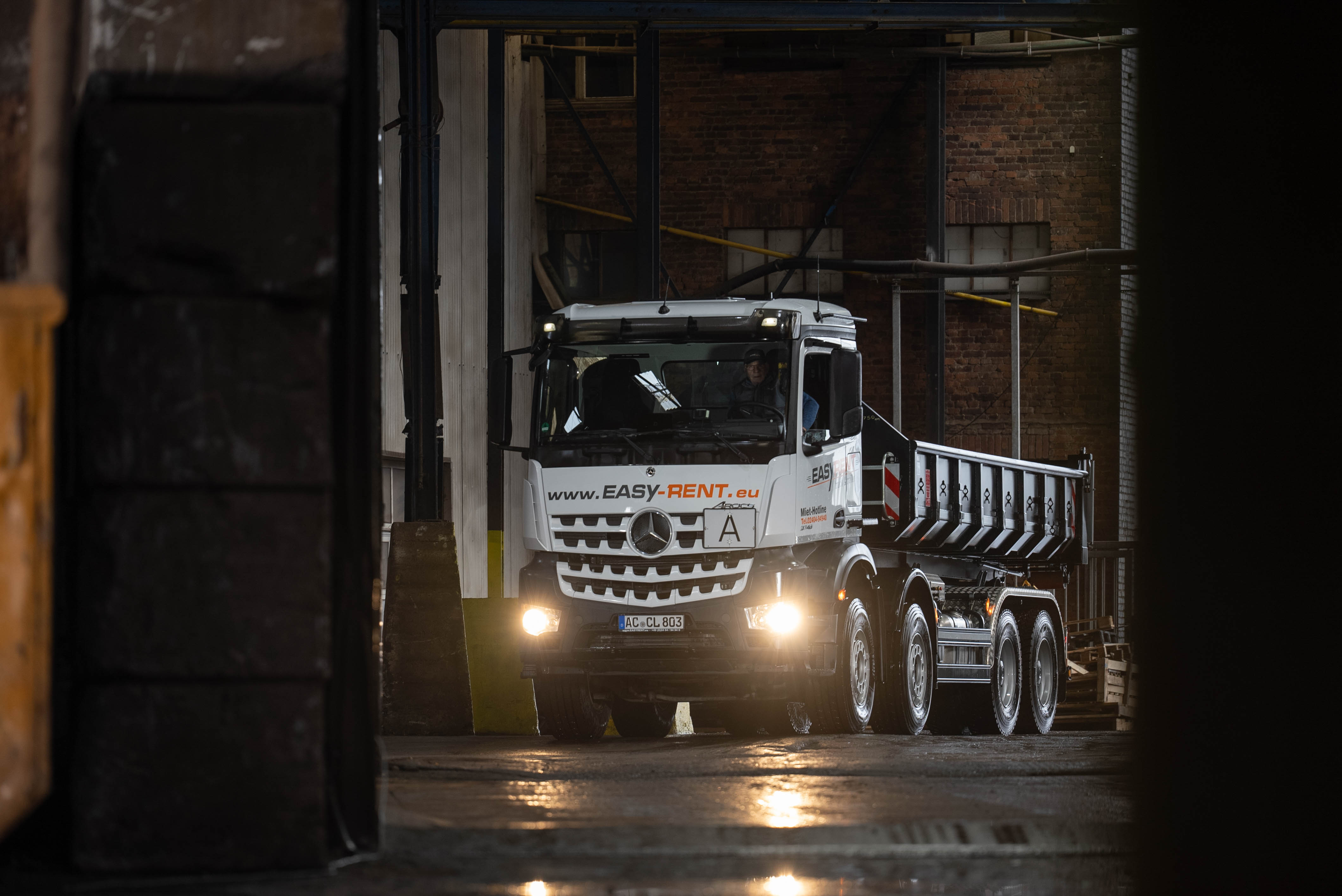 Witte Mercedes Benz 8x4 afrolkipper met container rijdt een afvalhal binnen