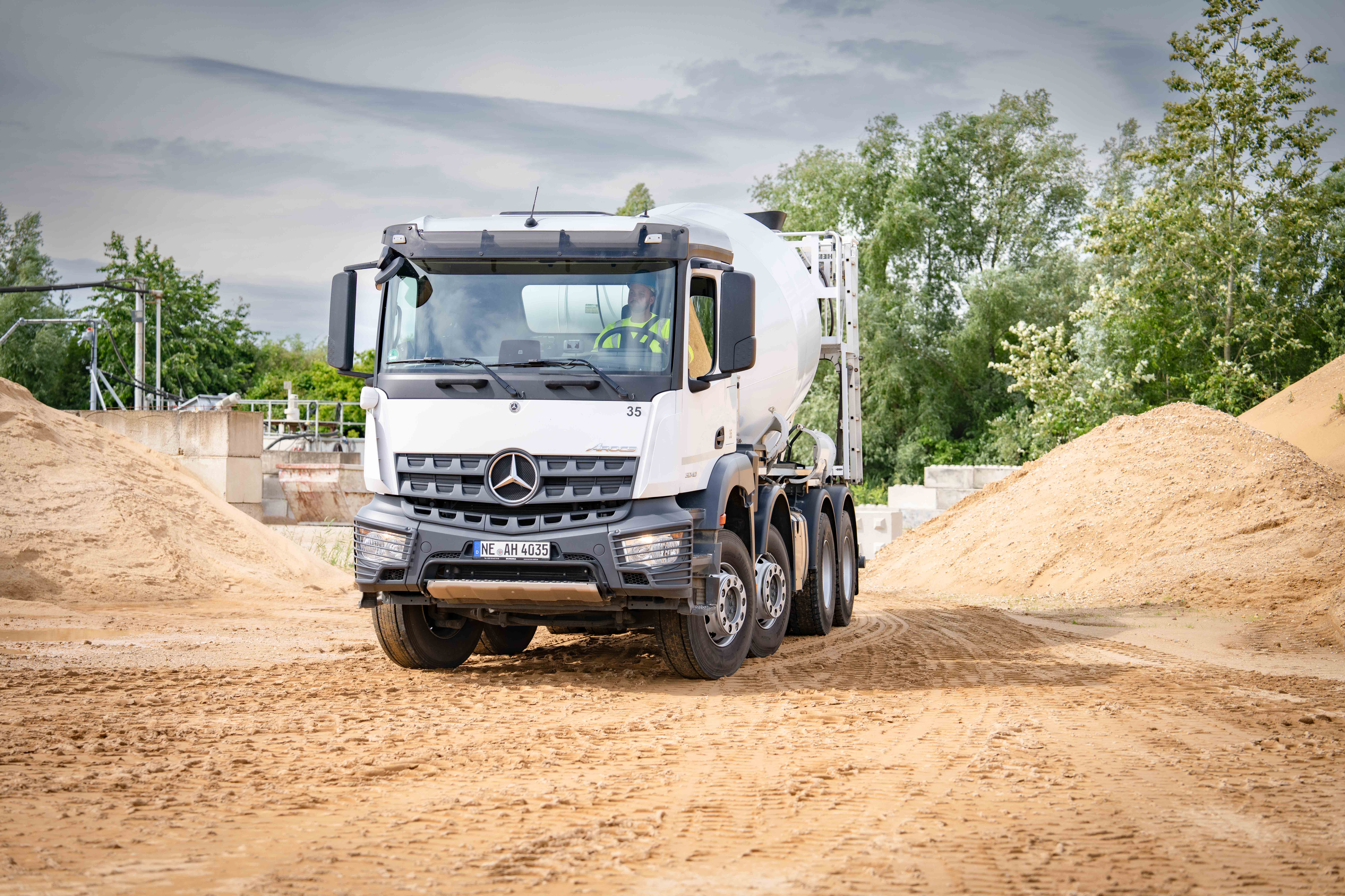 Weißer 8x4 Fahrbetonmischer von Mercedes-Benz Modell Arocs in einer Sandgrube, mit Bäumen im Hintergrund.