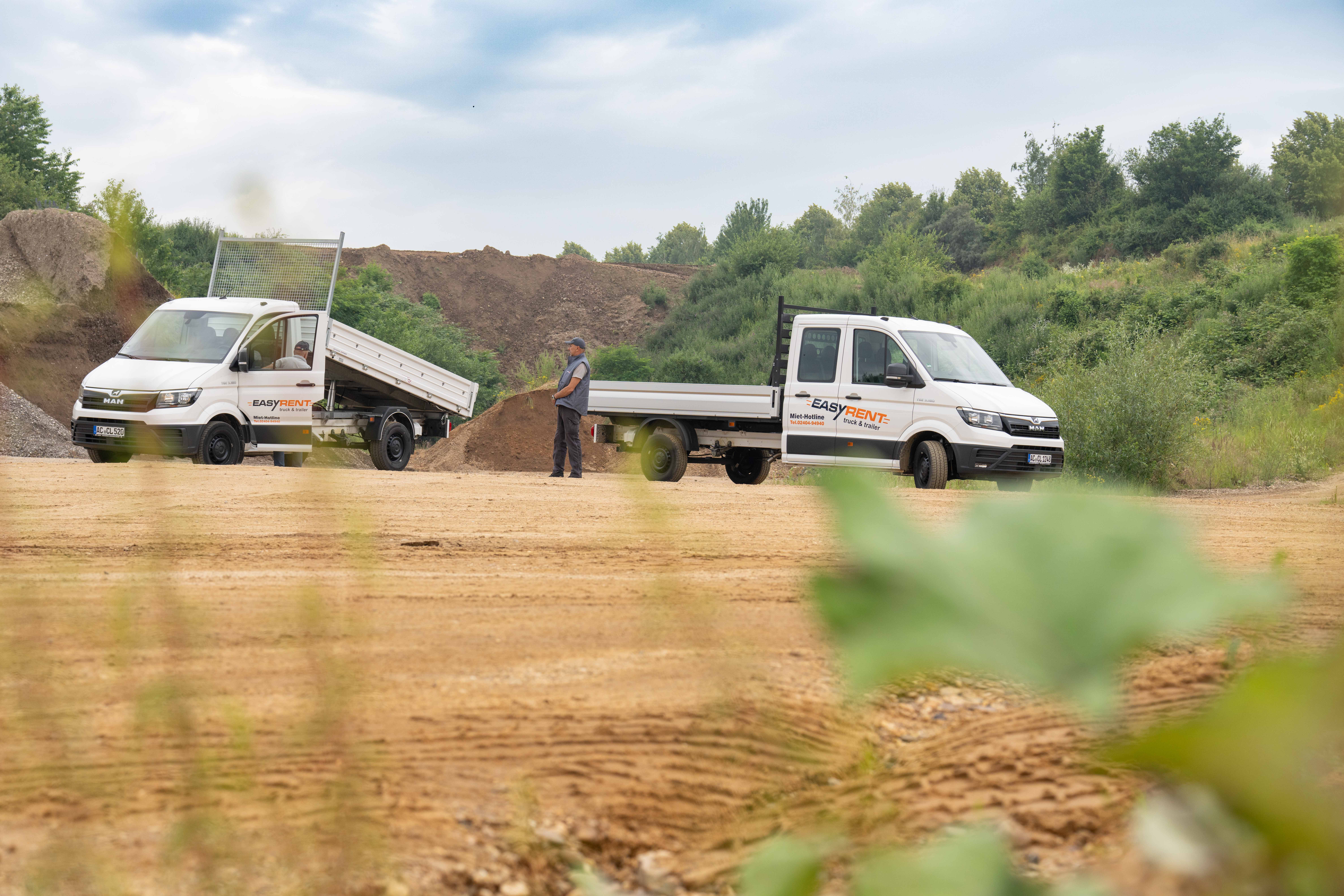 Zwei weiße MAN TGE 4x2 Transporter von Easy Rent auf einer Baustelle, einer mit gekippter Ladefläche. Ein Arbeiter steht zwischen den Fahrzeugen. Im Hintergrund sind Erdhügel zu sehen.
