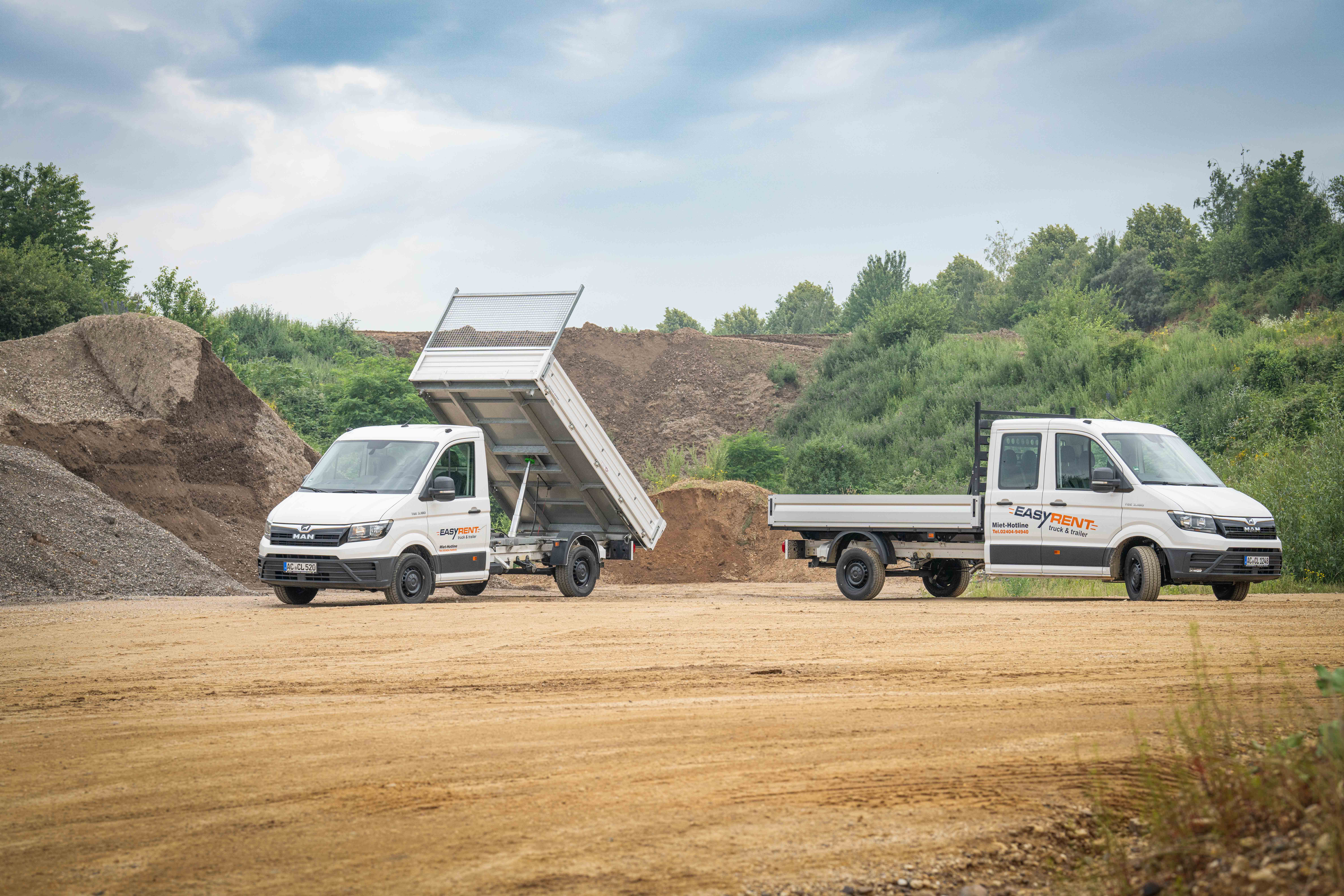 Zwei weiße MAN TGE 4x2 Transporter von Easy Rent auf einer Baustelle, einer mit gekippter Ladefläche beim Entladen von Schüttgut. Im Vordergrund ein Transporter mit der Aufschrift 'Easy Rent', im Hintergrund Sandhügel.
