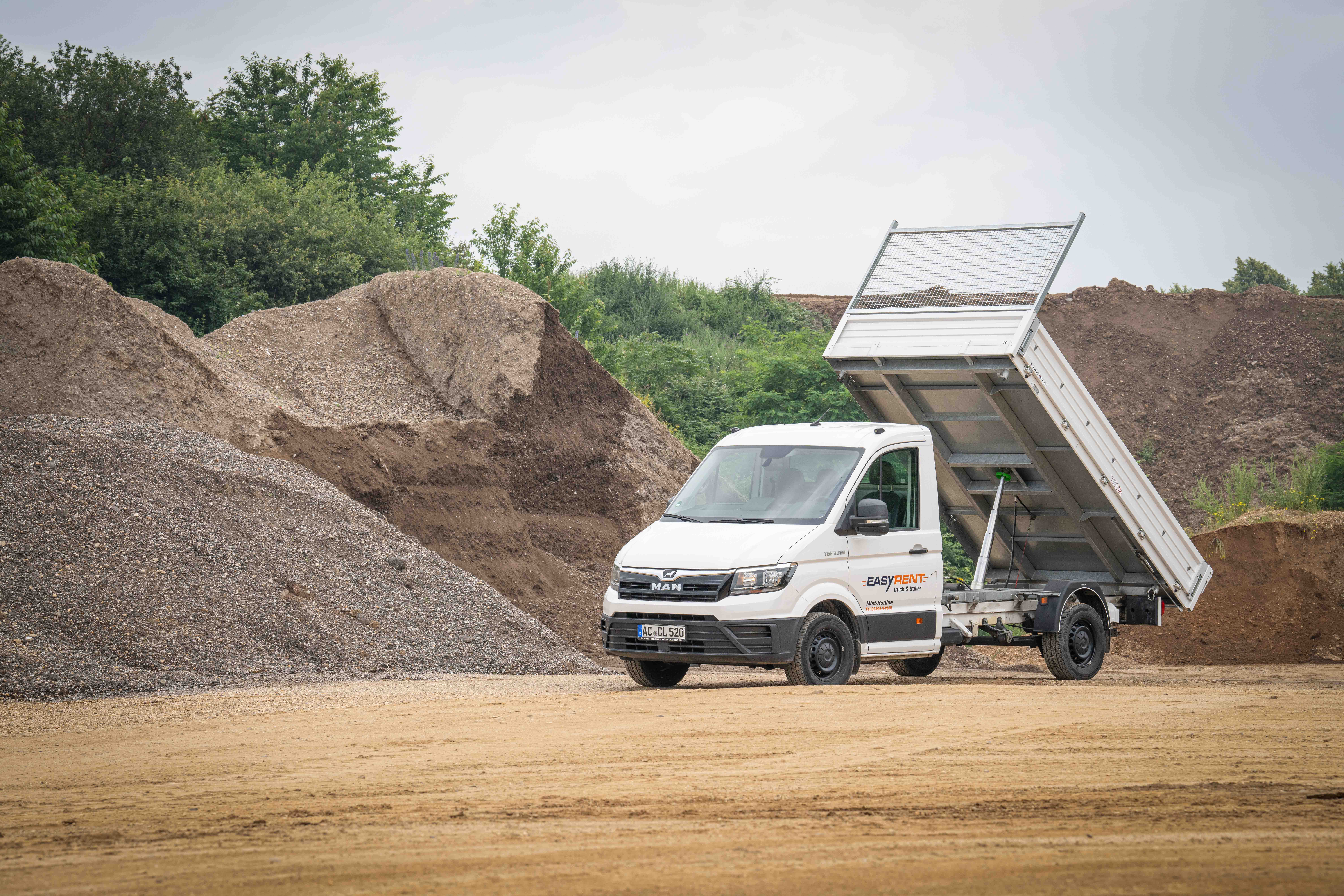 Weißer MAN TGE 4x2 Transporter Kipper von Easy Rent auf der Baustelle mit Kieshügeln. Die Ladefläche des Dreiseitenkippers ist nach hinten gekippt.