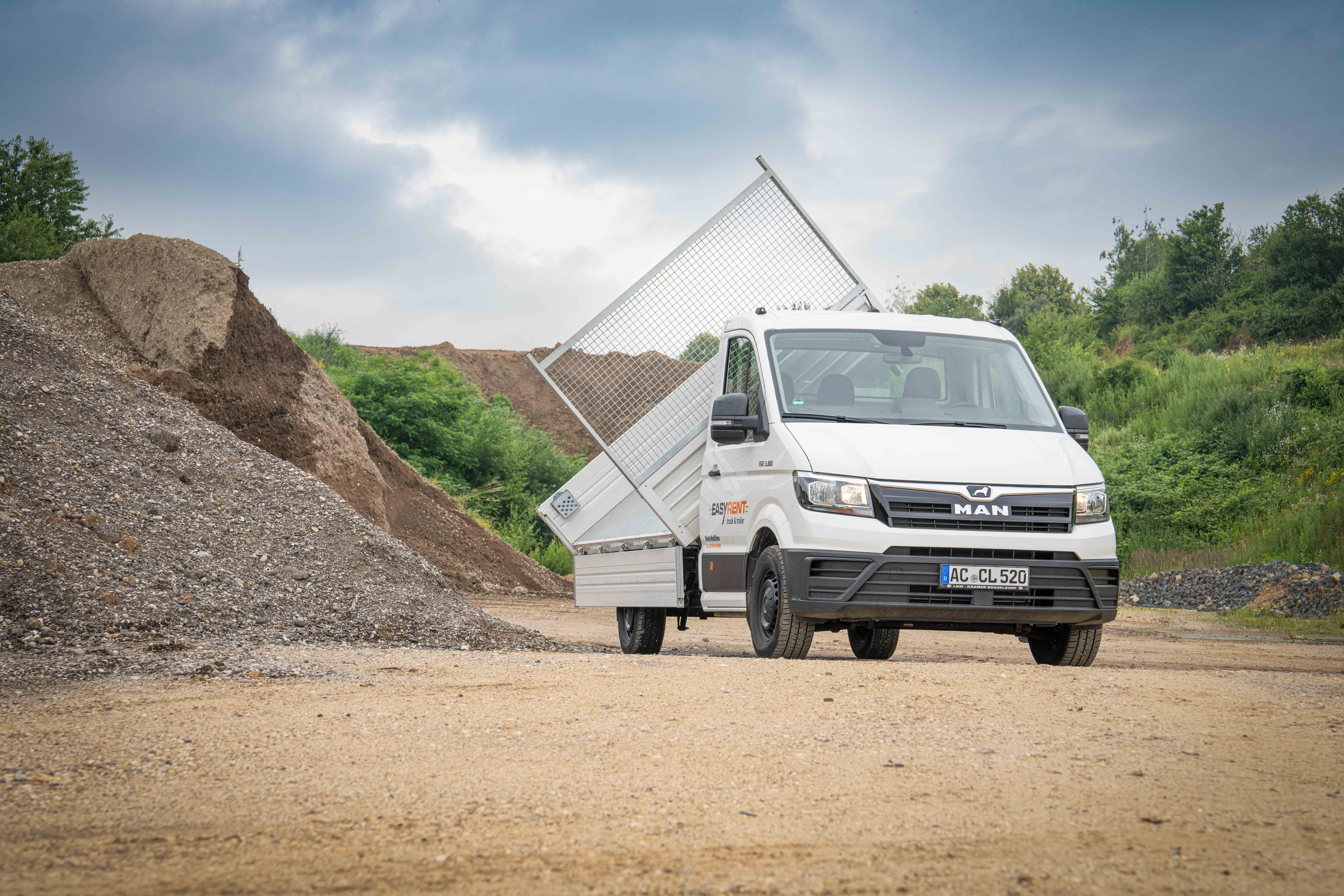 Ein weißer MAN TGE Dreiseitenkipper von Easy Rent auf einer Baustelle mit Sand- und Kieshügeln im Hintergrund. Die Ladefläche ist seitlich gekippt, was die Funktion des Fahrzeugs für gewerbliche Zwecke zeigt. Beschriftung 'Easy Rent' auf der Fahrertür.