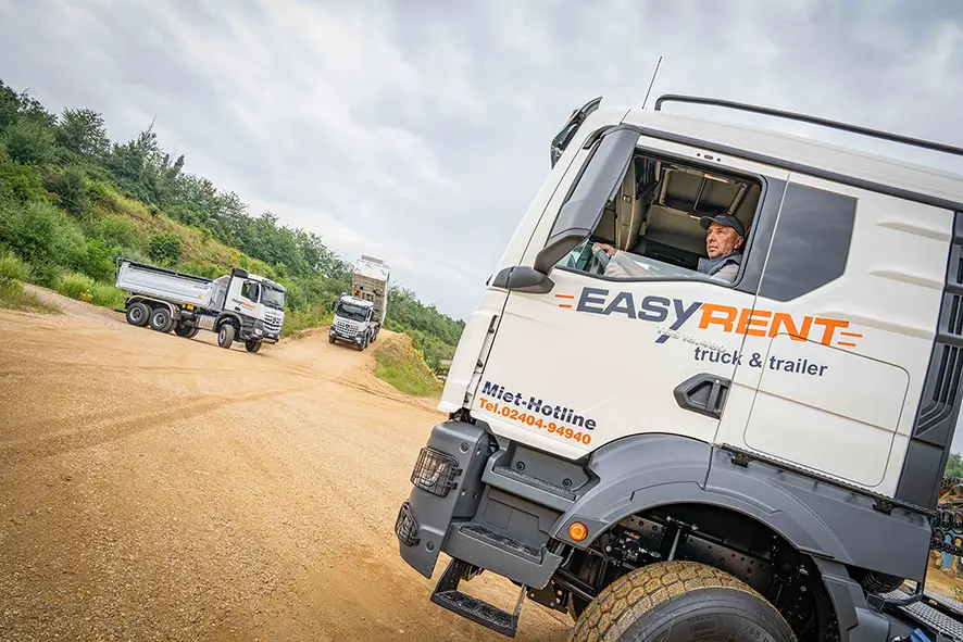 Camion-benne de EASY RENT en action sur une route de gravier. Au premier plan se trouve un camion MAN moderne portant l'inscription 'EASY RENT truck & trailer' et une hotline de location. Le chauffeur regarde attentivement depuis sa cabine. À l'arrière-plan, deux autres camions-bennes sont en action, dont un camion-benne arrière Mercedes à 4 essieux qui décharge des matériaux en vrac.