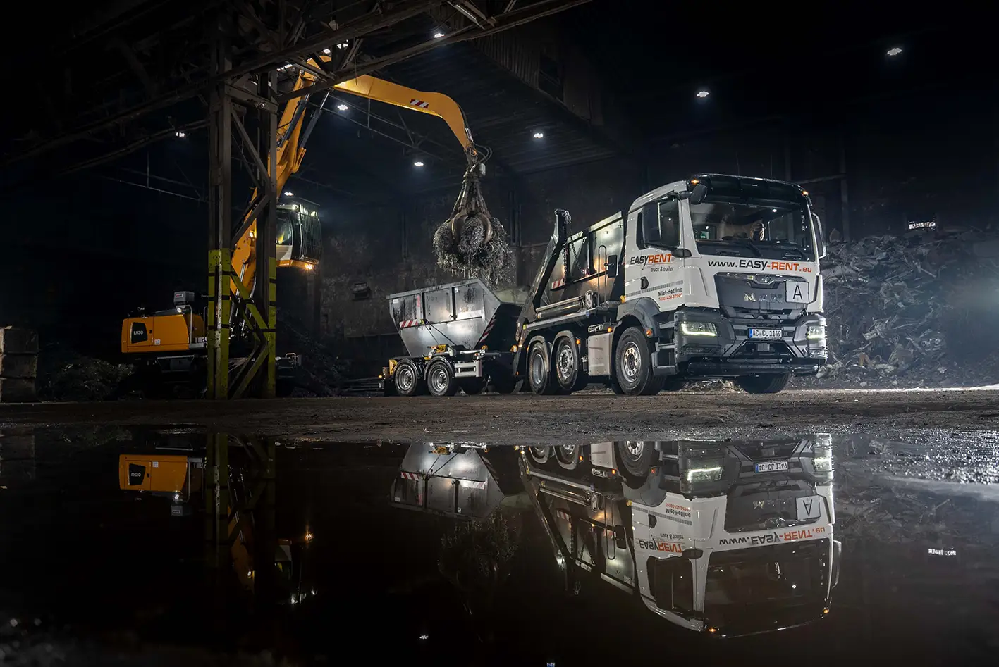 Ein MAN 3-Achs Absetzkipper steht in einer dunklen Industriehalle, während ein gelber Umschlagbagger Metallschrott in den Container lädt. Die Fahrzeugkombination besteht aus einem Absetzkipper mit Container, sowie einem 2-Achsanhänger mit Container.