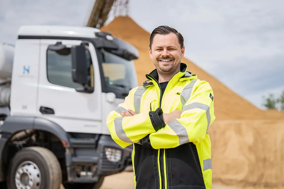 Pierre Wallenborn – Fuhrpark- und Standortleiter der Betonlogistik Deutschland im Rhein-Rhur-Gebiet steht in der Sandgrube und lächelt in die Kamera. Im Hintergrund steht ein Betonmischer.