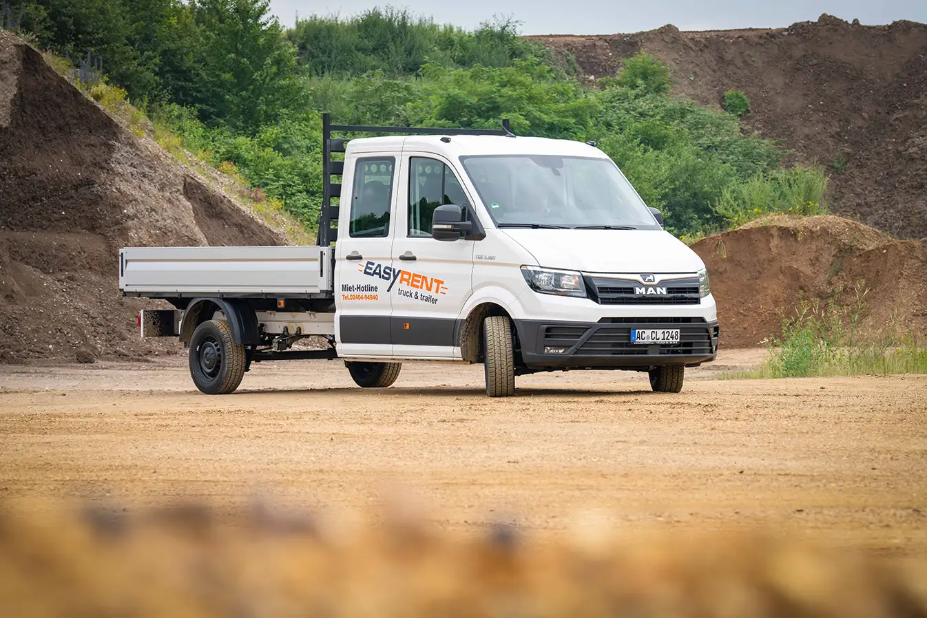 A white MAN TGE 4x2 transporter with a flatbed body from Easy Rent at a construction site. You can see hills of earth in the background. Ideal for commercial use in the construction industry.