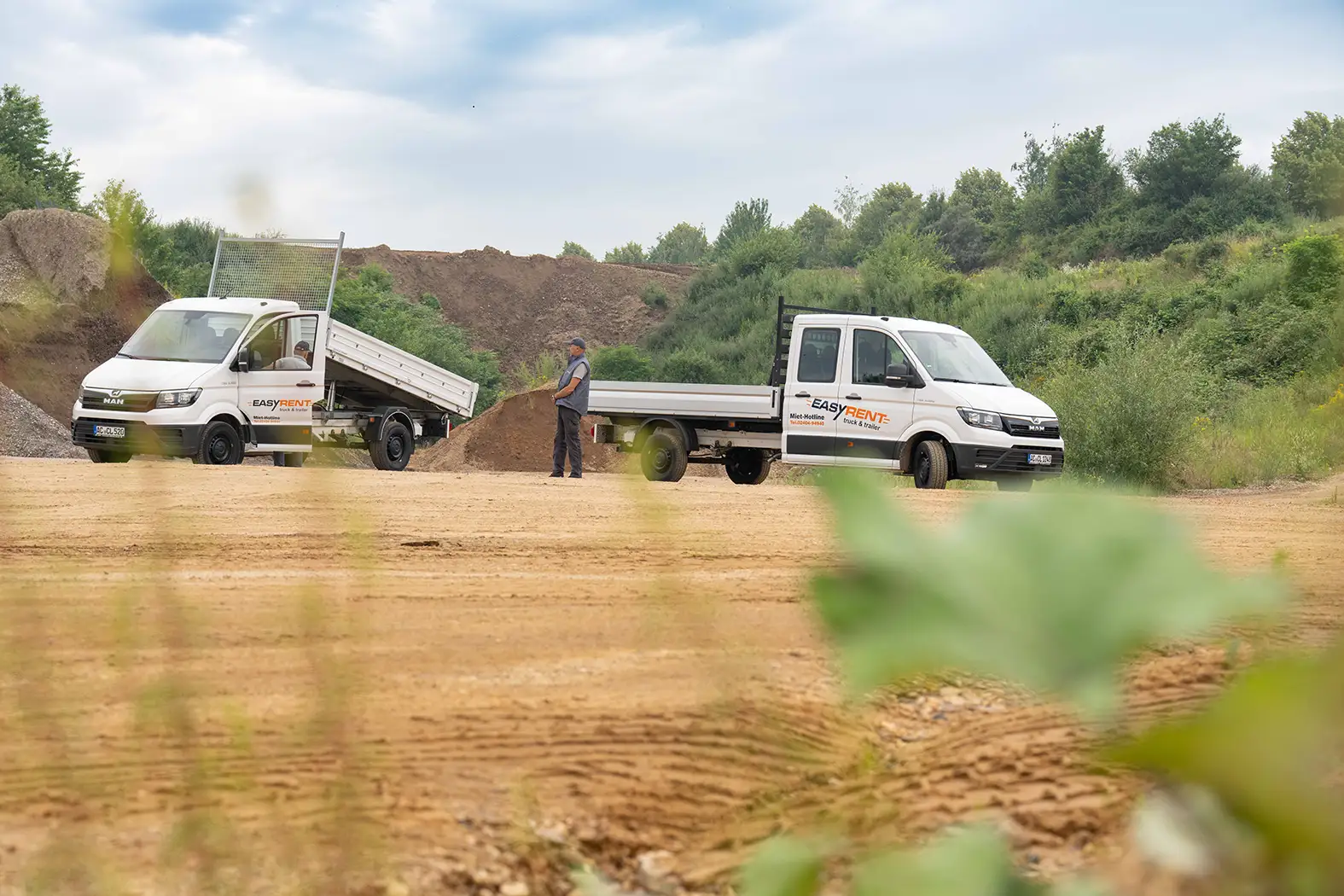 Two white MAN TGE 4x2 transporters from Easy Rent at a construction site, one with a tipped loading area. A worker stands between the vehicles. Mounds of earth can be seen in the background.