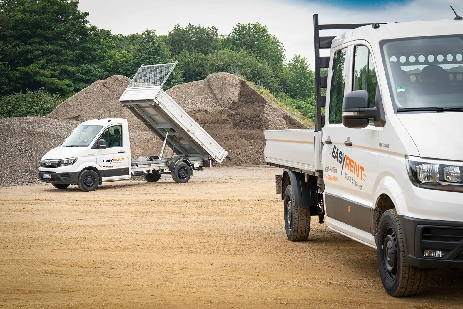 Two white MAN TGE 4x2 transporters from Easy Rent at a construction site, one with a tilted loading area unloading bulk material. In the foreground, a transporter with the inscription 'Easy Rent', in the background a sand hill.