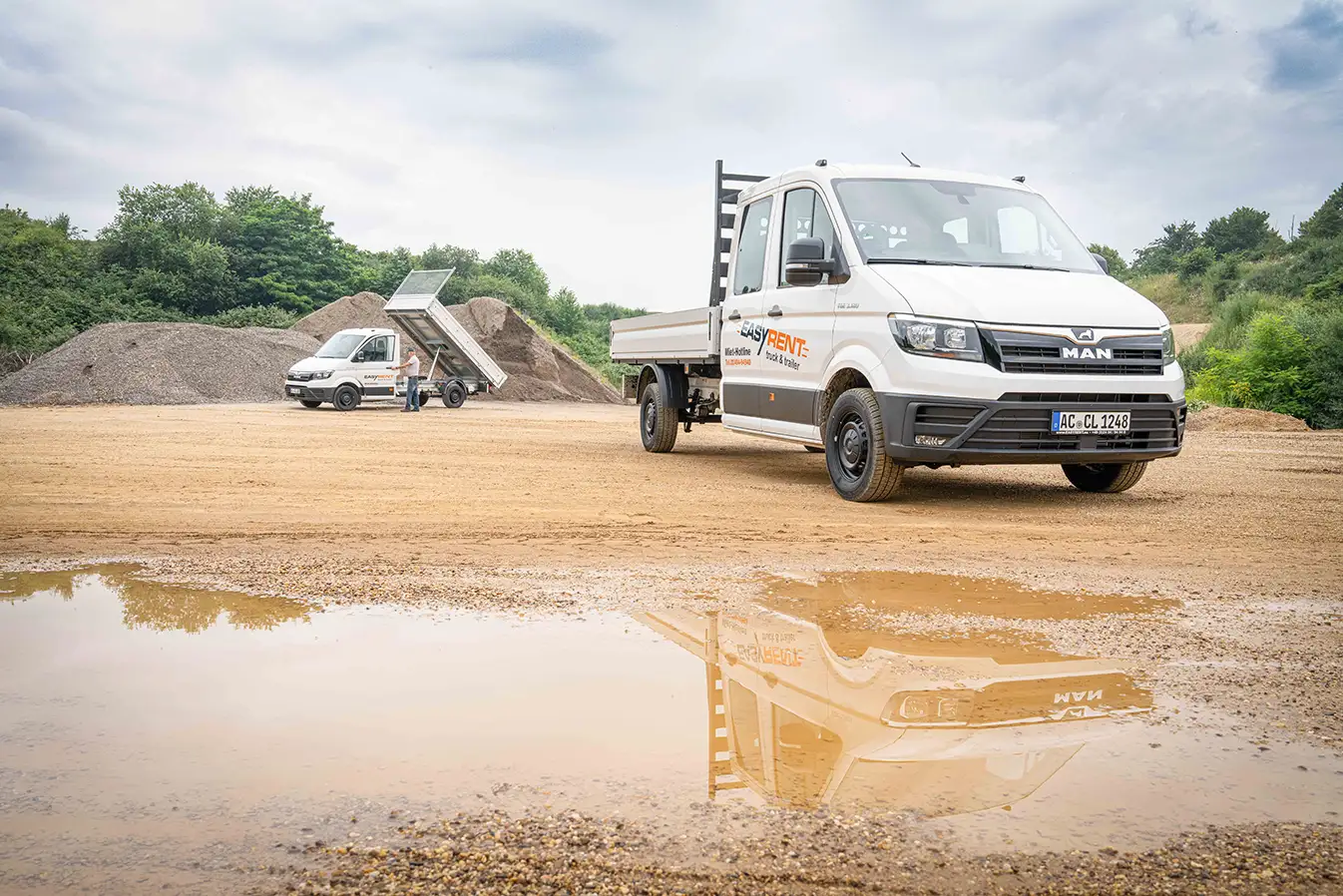 MAN TGE transporter from Easy Rent at a construction site. In the foreground, a transporter with a flatbed, reflected in a puddle; in the background, a sand hill and a second transporter tipper, with a tipped loading area.