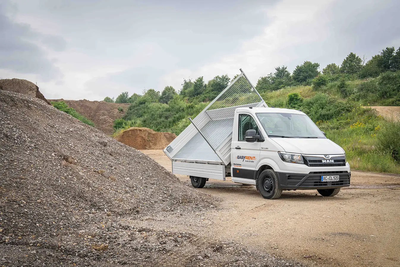 A white MAN TGE 4x2 three-way tipper transporter from Easy Rent on a construction site. The loading area is tipped to the side, ideal for transporting and unloading bulk materials. In the foreground is a pile of gravel.