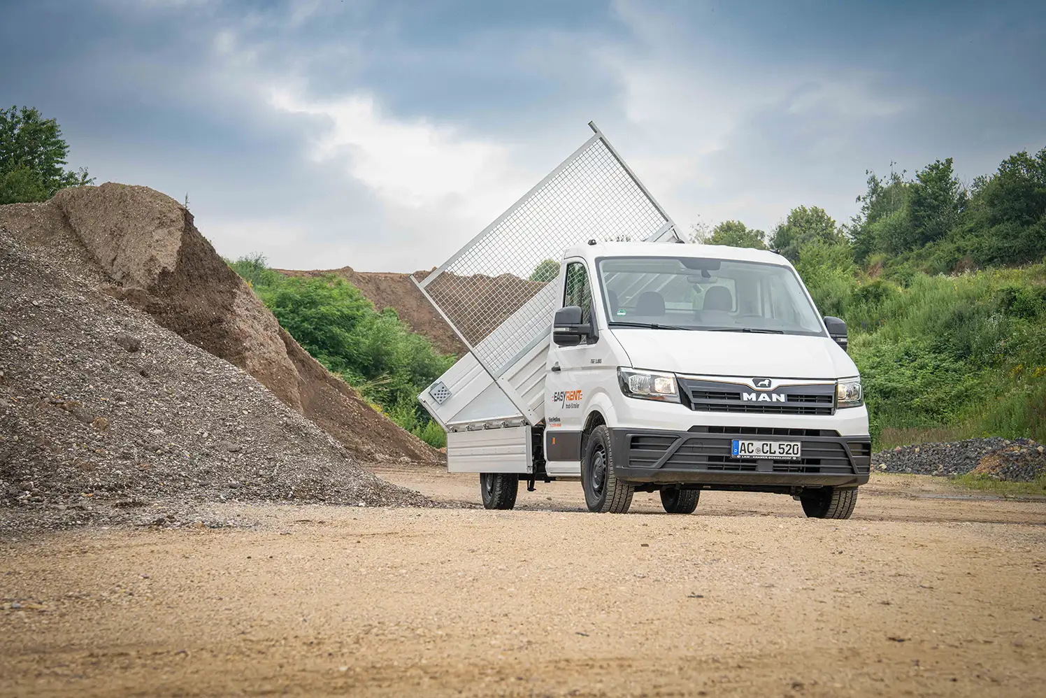 A white MAN TGE three-way tipper from Easy Rent at a construction site with sand and gravel heaps in the background. The loading area is tipped to the side, showing the vehicle's function for commercial purposes. 'Easy Rent' lettering on the driver's door.